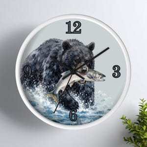 Puede incluir: Un reloj de pared redondo con marco blanco y esfera gris claro que muestra un oso negro atrapando un pez en el agua. El reloj tiene manecillas negras y números grandes de color gris oscuro en las posiciones 12, 3, 6 y 9.
