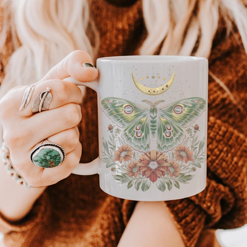 May include: White ceramic mug featuring a green and yellow moth design with a crescent moon and floral accents. The mug is held by a person wearing rings and a bracelet.
