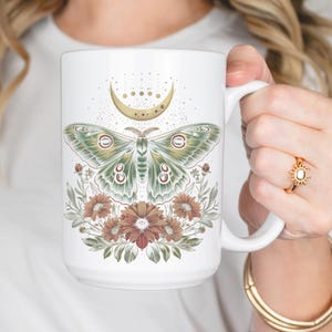 May include: White ceramic mug featuring a detailed illustration of a moth with green and cream wings, a gold crescent moon, and floral accents in shades of brown and green. The mug has a large handle and is held by a person.