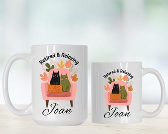 May include: White ceramic mug with a whimsical design featuring a pink armchair, two cats, and floral accents. The text "Retired & Relaxing" and the name "Joan" are printed on the mug. A smaller matching mug is also present.