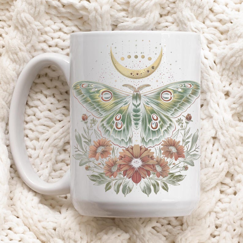 May include: White ceramic mug featuring a detailed illustration of a green and cream moth with a crescent moon and floral accents. The mug has a large handle and is decorated with orange and red flowers.