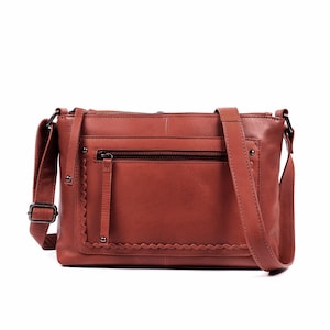 May include: A brown leather crossbody bag with a long adjustable strap. The bag features a front zippered pocket with a scalloped edge and a main compartment with a zipper closure. The bag has a rectangular shape and is made of a smooth leather material.