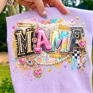 May include: A light purple t-shirt featuring the word MAMA in large, decorative letters. The design includes gold glitter, pink and blue tie-dye patterns, pearl accents, dangling heart-shaped charms, a safety pin, and small colourful rhinestones.