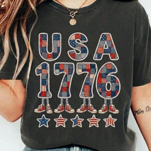 May include: A charcoal grey t-shirt featuring the text USA 1776 in a patchwork pattern of red, white, and blue stars and stripes. Each character has legs wearing red trainers. Below the text are five stars with red, white, and blue patterns.
