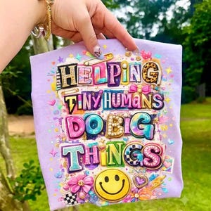 May include: A light purple t-shirt featuring a colorful graphic with the text "HELPING TINY HUMANS DO BIG THINGS" in various fonts and patterns. The design includes a yellow smiley face, small hearts, stars, bows, and a safety pin illustration.