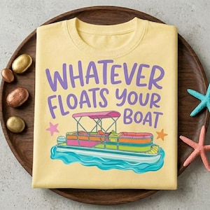 May include: A pale yellow t-shirt featuring a graphic of a colourful pontoon boat on blue water. Above the boat, the text reads "WHATEVER FLOATS YOUR BOAT" in purple letters. Two small pink and orange stars are positioned on either side of the boat.