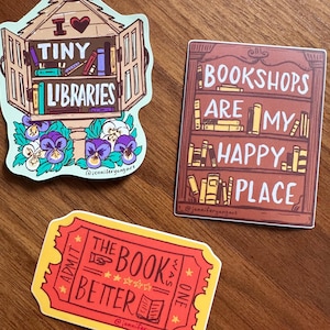 May include: Three stickers on a wooden surface. One sticker is a tiny library with the text "I Tiny Libraries". Another sticker says "Bookshops are my happy place". The third sticker is a red ticket that says "The book was better".