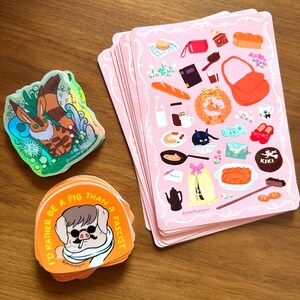 Pu&ograve; includere: Una collezione di adesivi e biglietti illustrati. Biglietti rosa con cibo e accessori, adesivi arancioni con un design a maiale e testo, e un adesivo olografico con un animale dei cartoni animati sono visibili.