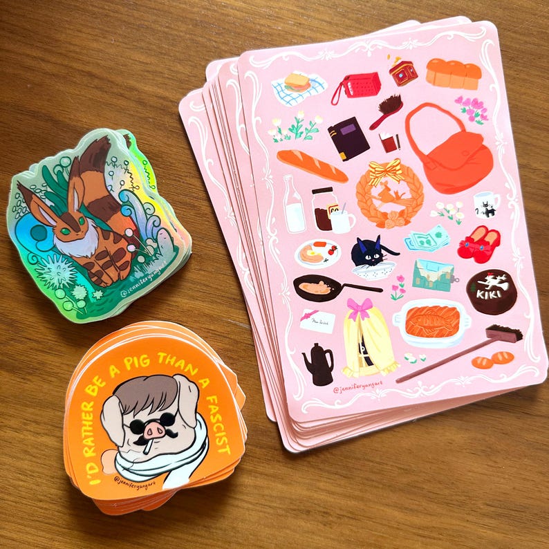 Pu&ograve; includere: Una collezione di adesivi e biglietti illustrati. Biglietti rosa con cibo e accessori, adesivi arancioni con un design a maiale e testo, e un adesivo olografico con un animale dei cartoni animati sono visibili.