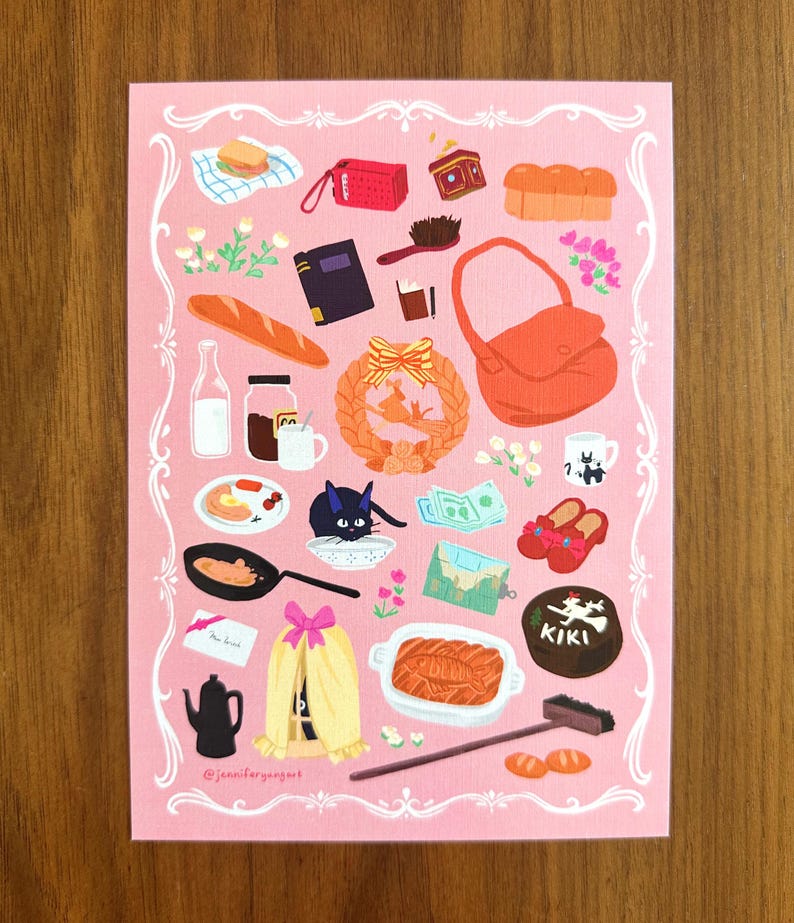 May include: Pink art print featuring various illustrated objects, including a loaf of bread, a purse, a black cat, and a teapot. Other items include a book, a frying pan, and a cake with the word "KIKI" on it. The print has a decorative border.