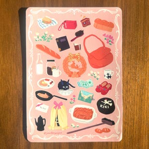 Pu&ograve; includere: Illustrazione rosa con cornice decorativa, raffigurante vari oggetti, tra cui una borsa rossa, un gatto nero e una baguette. L'illustrazione include il testo "KIKI" e "@jennifergangkuk".