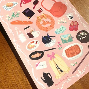 Pu&ograve; includere: Un foglio di adesivi rosa con illustrazioni di un gatto nero, una teiera, una borsa e cibi come pane e un uovo fritto. Il foglio ha un bordo decorativo e il testo "New Witch".