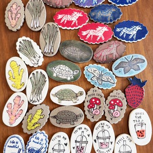 May include: A collection of hand-painted ceramic pieces with various designs. The designs include horses, turtles, butterflies, ginger, scallions, mushrooms, strawberries, bubble tea, and noodle bowls. The pieces are in various colors and shapes, with some featuring text.