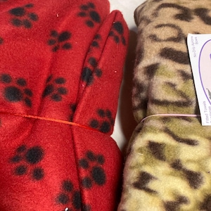 May include: Close-up of two fleece items. One is bright red with black paw prints, tied with orange string. The other is tan and brown with a leopard print. The items appear to be folded or bundled.