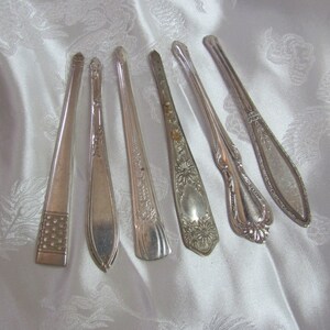 May include: Six silver-plated serving spoons with ornate handles. The spoons have different designs and are arranged in a row on a white satin fabric.