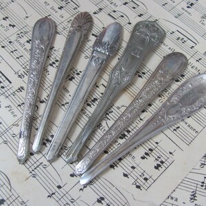 May include: Six silver spoons with ornate designs, arranged in a fan shape on a sheet of music. The spoons have a polished finish and intricate details.