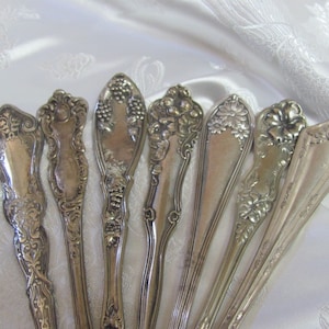 May include: Seven silver spoons with ornate floral and grape designs. The spoons are arranged in a fan shape on a white fabric background.