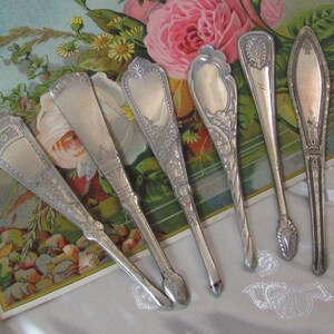 May include: Six antique silver butter knives with ornate designs. The knives are arranged on a floral background with a vintage aesthetic. Each knife features a unique handle and blade shape, showcasing intricate details.