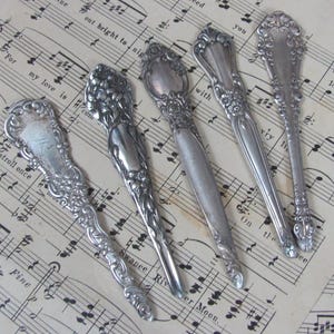 May include: Five antique silver spoons with ornate designs, arranged on a sheet of music. The spoons have a vintage look and are perfect for adding a touch of elegance to any table setting.