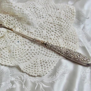 Peut inclure: Un crochet vintage avec un manche argenté orné et un petit crochet incurvé. Le crochet repose sur un napperon blanc et dentelle, suggérant un thème artisanal. La poignée présente des volutes détaillées.