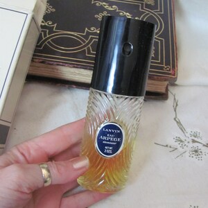May include: A vintage perfume bottle with a black cap and a clear, textured glass body. The bottle has a blue and white label that reads "LANVIN EAU ARPEGE atomiseur" and contains a yellow liquid. The bottle is 2 oz.