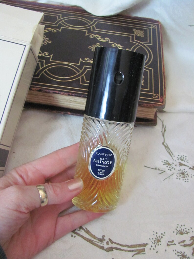 May include: A vintage perfume bottle with a black cap and a clear, textured glass body. The bottle has a blue and white label that reads "LANVIN EAU ARPEGE atomiseur" and contains a yellow liquid. The bottle is 2 oz.
