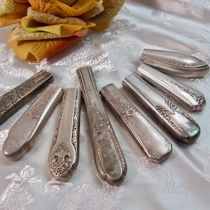 Könnte beinhalten: Eine Sammlung von sieben silberfarbenen Messergriffen aus Metall mit verschiedenen geprägten Blumen- und geometrischen Mustern. Jeder Griff ist länglich mit einem abgerundeten Ende und weist eine metallische Oberfläche mit Altersspuren und Patina auf.