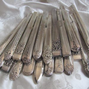 May include: A collection of antique silver silverware, including forks, knives, and spoons. The silverware features ornate floral and geometric designs. The pieces are arranged on a white, textured surface, showcasing their polished, reflective surfaces.