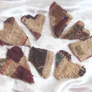 May include: A selection of handmade patchwork heart-shaped decorations. Each heart is crafted from various textured fabrics in beige, brown, and burgundy, with visible stitching and frayed edges. The hearts are displayed on a white, patterned background.