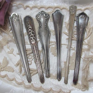May include: Six silver-plated, ornate, decorative spoons with various designs. The spoons are arranged in a row on a white lace tablecloth.