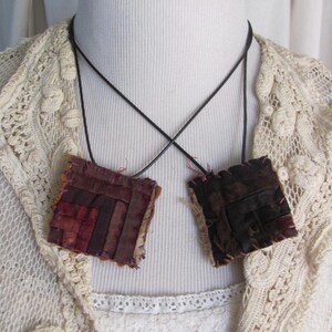 May include: A handmade necklace with two square pendants made of layered fabric scraps in shades of brown, burgundy, and cream. The pendants are attached to a black cord.