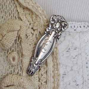 Op de afbeelding: Een zilverkleurige antieke broche met sierlijke details en een gegraveerde initiaal "E". De broche is geplaatst tegen een achtergrond van crèmekleurig kant en haakwerk.