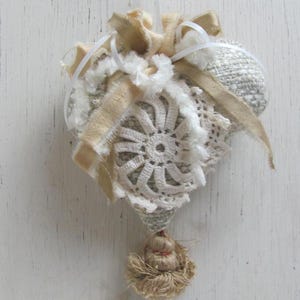 May include: A heart-shaped ornament made of beige fabric with white lace and a brown tassel. The ornament is decorated with white and gold ribbon.