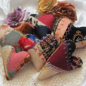 以下が含まれることがあります： 赤、青、黒、白など、さまざまな色とパターンで手作りされたパッチワークのハート型のクッションの集まりです。クッションは、複雑な刺繍で飾られており、白いレースのテーブルクロスの上に置かれています。