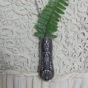 May include: A silver spoon necklace with a fern sprig in the spoon bowl. The necklace is worn on a white lace top.