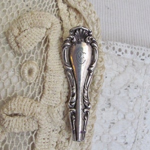May include: A silver-toned, ornate, decorative spoon with a floral design and a monogrammed letter "C" on the handle. The spoon is resting on a white crocheted lace fabric.