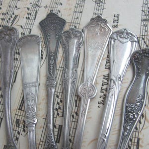 Op de afbeelding: Een assortiment antieke zilveren serveerbestekken, waaronder lepels en vorken, met sierlijke bloemen- en geometrische ontwerpen. Het bestek is gerangschikt op een achtergrond van vintage bladmuziek, wat een nostalgische uitstraling creëert.