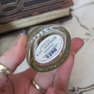May include: A clear glass perfume bottle with a white label that reads "LANVIN EAU ARPEGE atomiseur." The bottle is held in a hand, with a gold ring visible. The label also includes the number "4288."