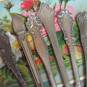 May include: Five antique silver spoons with ornate designs. The spoons are arranged on a floral patterned surface with pink roses.