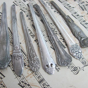May include: An assortment of antique silver spoons and serving utensils with ornate floral and scroll designs. The spoons are arranged on a sheet of vintage sheet music, showcasing their intricate details and aged patina.