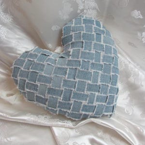 May include: A heart-shaped pillow made from woven denim patches. The pillow has a light blue color with white stitching and frayed edges, creating a textured appearance. The pillow is a decorative item.