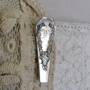 May include: A silver-toned coffin-shaped brooch with an ornate design and the letter "F" in the centre.