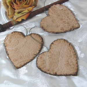 May include: Three heart-shaped ornaments made of light tan quilted fabric with visible stitching patterns. Each heart features dark, burnt-looking edges and a thin grey string attached at the top for hanging. The hearts have a textured surface.
