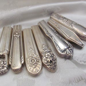 Puede incluir: Una colección de mangos de cubiertos antiguos plateados, que muestran varios diseños florales y ornamentados. Los mangos están dispuestos sobre una superficie blanca texturizada, resaltando los intrincados detalles y la pátina envejecida de la platería.