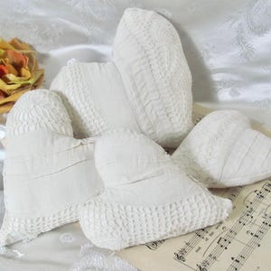 May include: A collection of four off-white heart-shaped fabric ornaments featuring various textures including smooth cotton, pleated fabric, and intricate crochet lace detailing. The hearts are arranged together, showcasing different sizes and patterns.