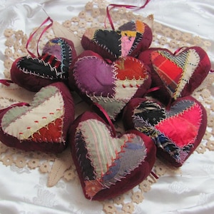 以下が含まれることがあります： 赤、ピンク、白の色の8つの手作りパッチワークハート。各ハートは異なる生地で作られており、装飾的なステッチで縫い合わされています。ハートは白いレースのテーブルクロスから吊り下げられています。