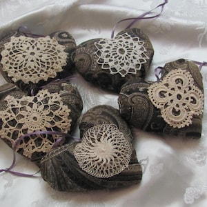 以下が含まれることがあります： 白い布の表面に、レースのドイリーが付いた茶色の布製のハート型のクッションが6つ、半円形に並んでいます。