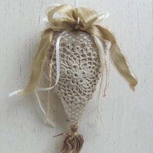 May include: A beige crocheted heart-shaped ornament with a gold ribbon bow and a gold tassel. The ornament is hanging from a white string.