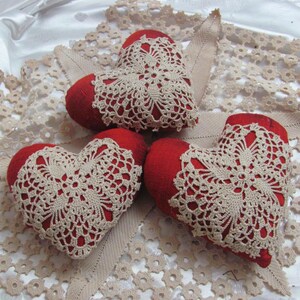 以下が含まれることがあります： 白いレースのテーブルクロスの上に、赤いフェルトのハート型のクッションが3つ置かれ、その上にクロシェレースのドイリーが置かれています。