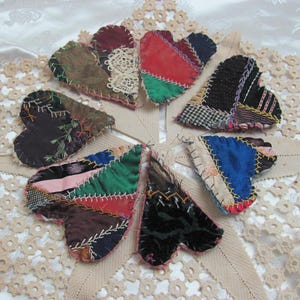 Puede incluir: Ocho corazones de patchwork en varios colores y patrones, incluyendo rojo, verde, azul, marrón y negro. Los corazones están dispuestos en un círculo sobre un mantel de encaje blanco.